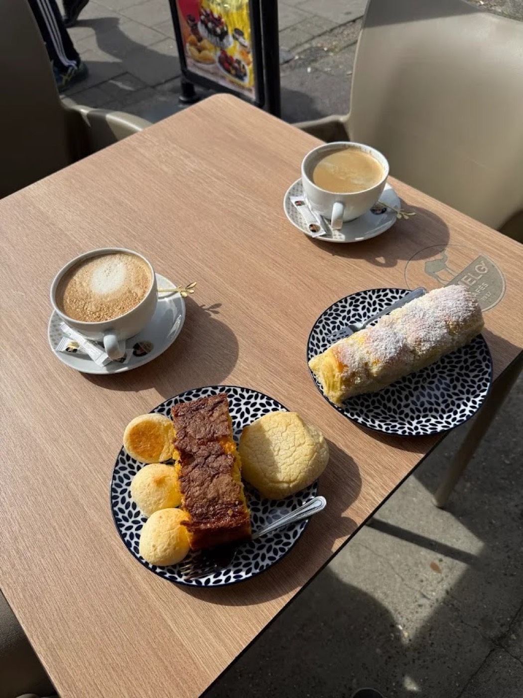 Two coffees with p&atilde;o de queijo, a slice of cake and a sugar-dusted roll on a sunny table